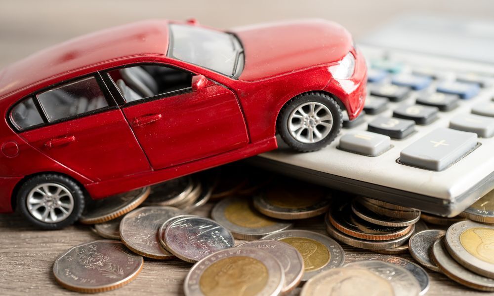Imagen de cerca de un coche rojo de juguete sobre una mesa de madera apoyado en una calculadora gris y varias monedas de dos euros.