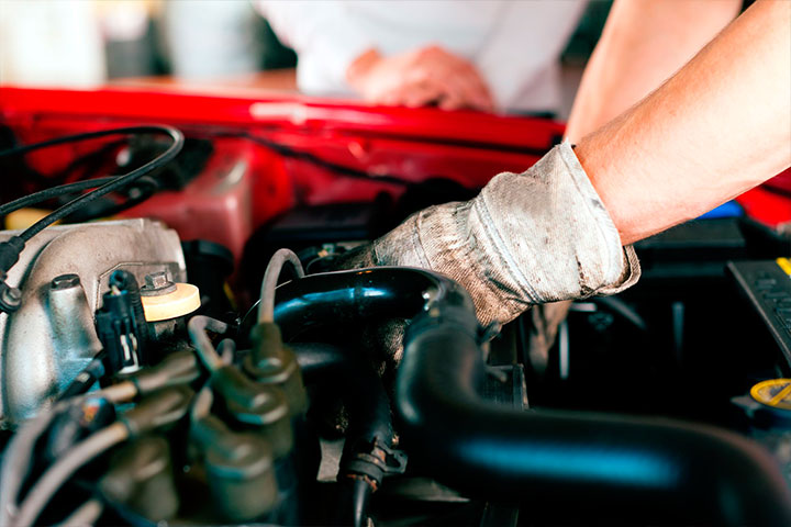 Fotografía en plano de detalle de la mano de un mecánico sobre el motor de un coche de color rojo, intentando reparar una avería mécanica. | Mycaready