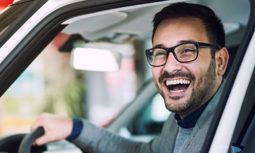 Imagen de cerca de un conductor feliz y sonriente sentado en un coche blanco a punto de arrancar con el dedo pulgar levantado haciendo el signo de okey.