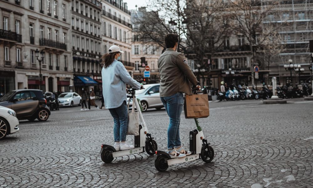 Foto de un hombre y una mujer circulando con dos patinetes eléctricos por el centro de la ciudad en pareja.