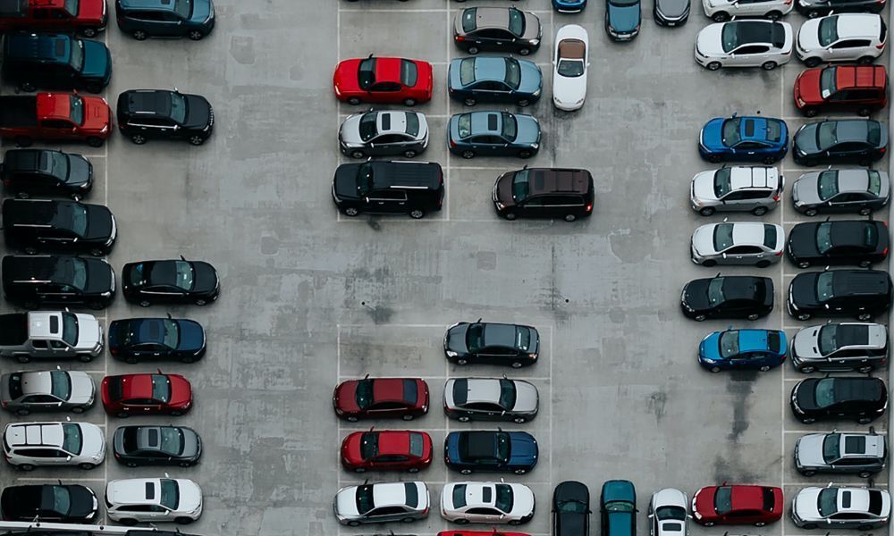 Imagen aérea desde arriba de un aparcamiento de asfalto de color gris lleno de distintos modelos de coches actuales y de colores varios, la gran mayoría aparcados.