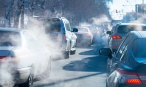 Imagen de cerca de varios coches circulando por la carretera en el medio de la ciudad, y emitiendo en exceso varios gases contaminantes para el medio ambiente.