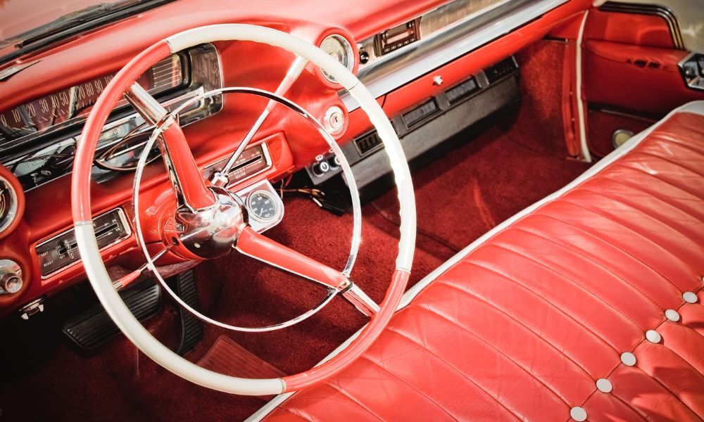 Interior de un coche clásico de color rojo con detalle en el volante blanco y en la parte delantera del asiento del piloto y el copiloto.