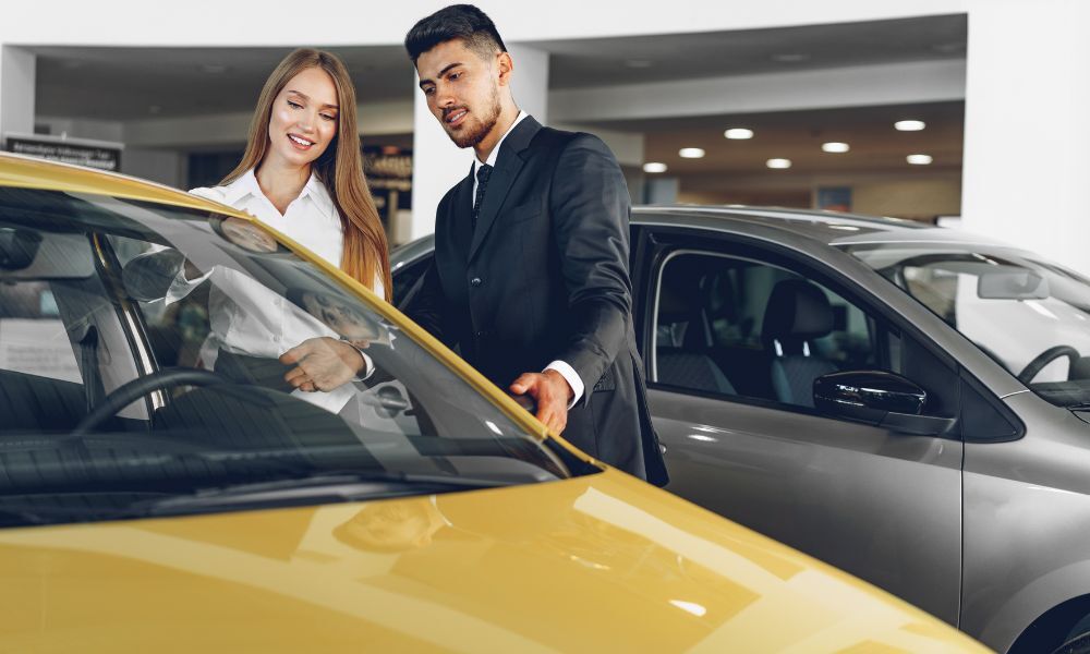 Vendedor en concesionario de coches de segunda mano, enseñando un vehículo compacto de color amarillo a una mujer joven que lo quiere comprar.